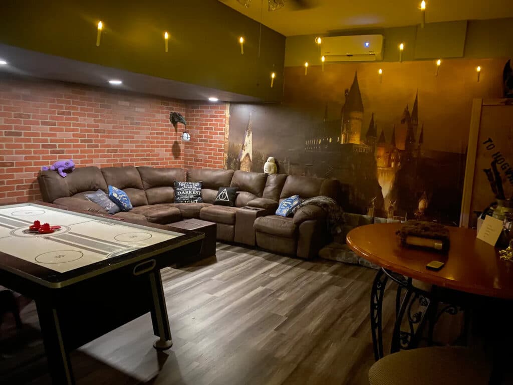A dimly lit living room with bricked walls and wooden flooring. Suspended lights mimic floating candles while a mural of Hogwarts is on the focus wall.